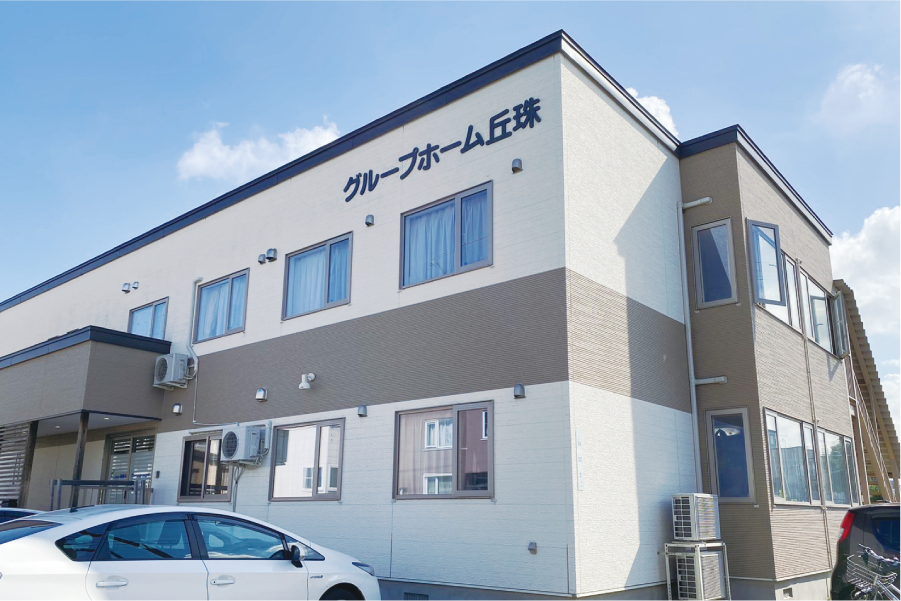 施設はホワイトとライトグレーの柔らかな雰囲気の２階建ての建物である。入口には階段と手すり付きのスロープがある。