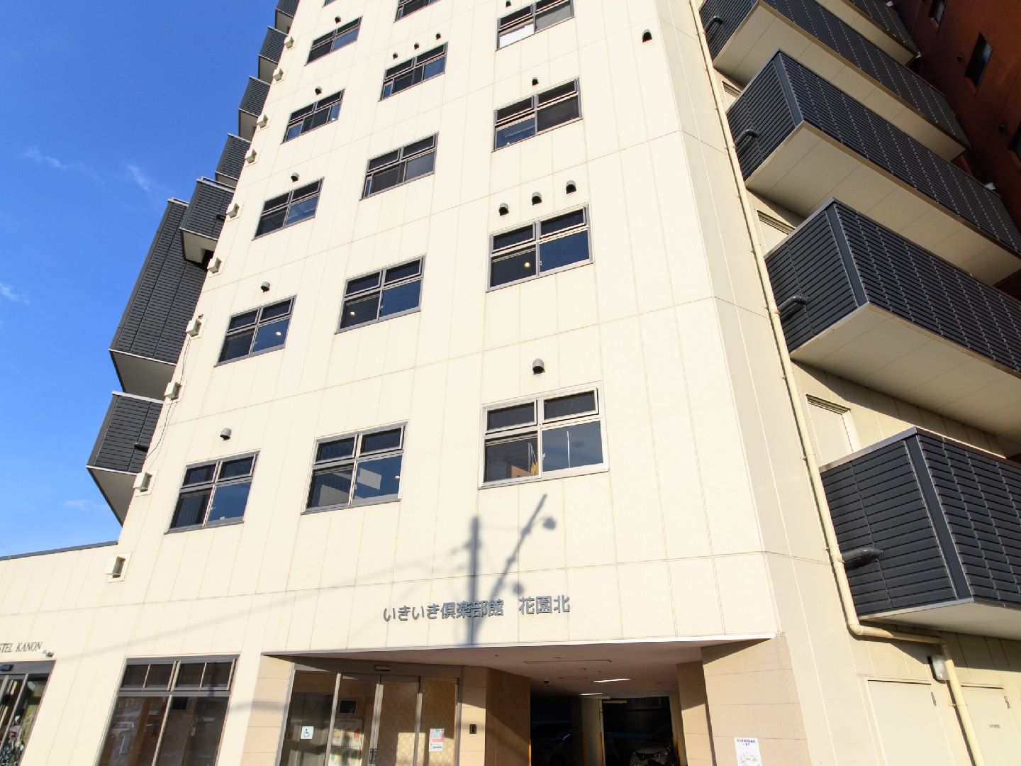 施設は大きな通り沿いに立地している。白い壁と黒のバルコニーがモダンな雰囲気の11階建ての施設の外観。1階には駐車場スペースがある。
