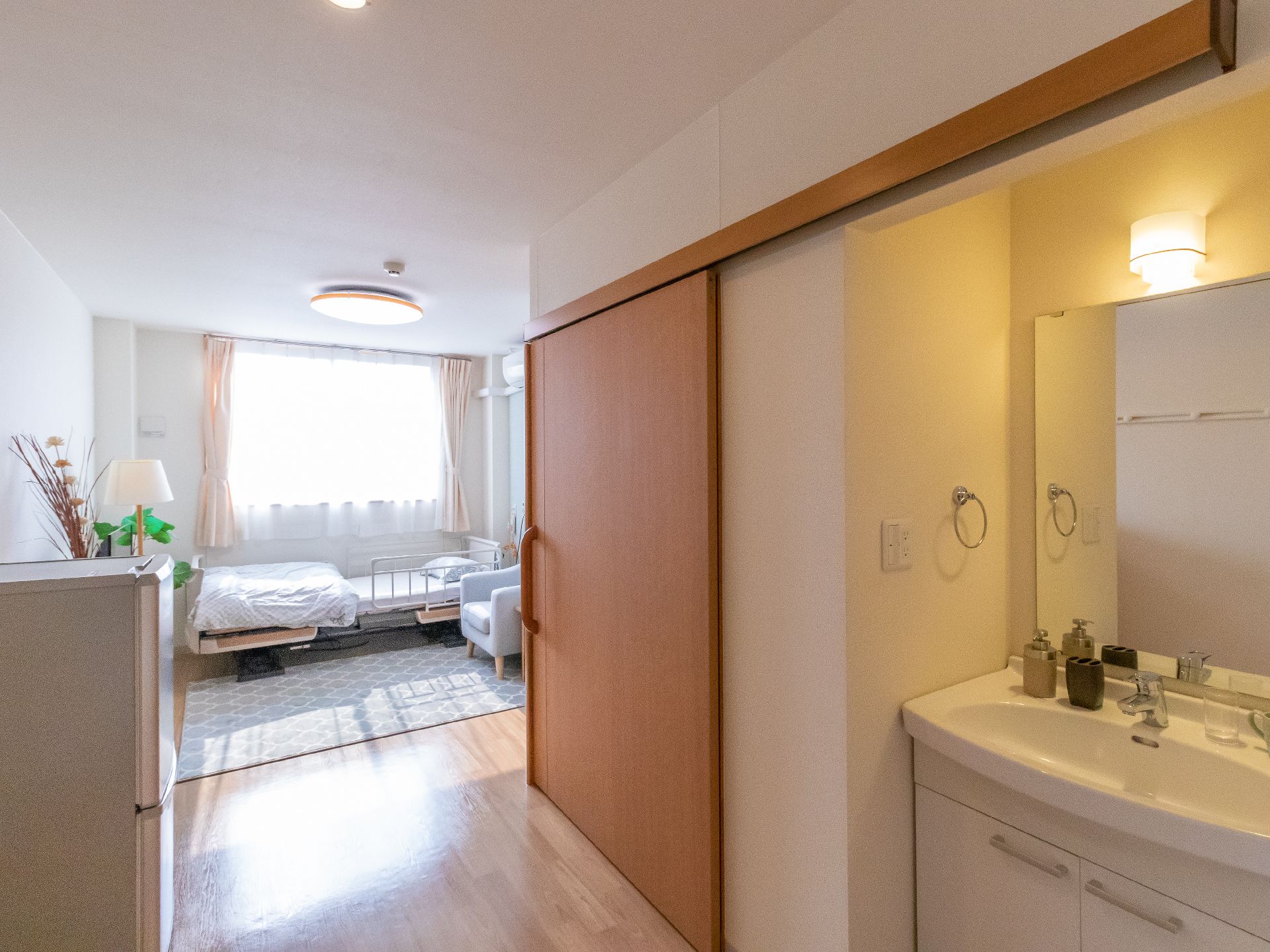奥に大きな窓が付いたベッドの置いてある部屋がある洗面台が置かれた通路