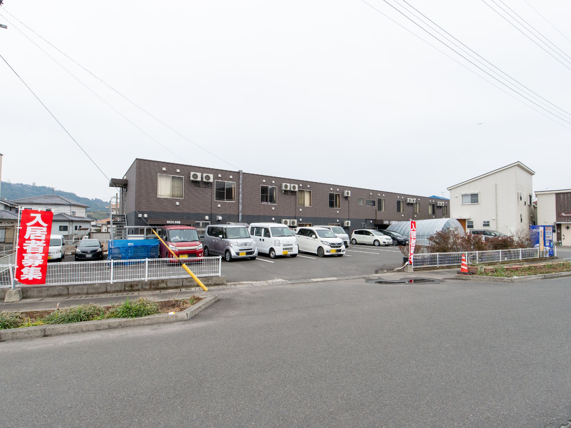 広い道路に面したグループホームで、敷地内にアスファルトで舗装された駐車場が完備されている。周辺には高い建物がない。