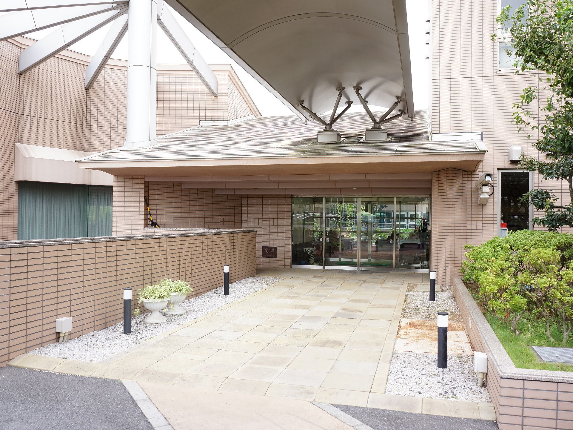 施設のエントランス。入口までの通路両脇に、小石を敷き詰めたスペースがあり、照明や鉢植えの植物がある。上方に、アーチ状の屋根が付いている。