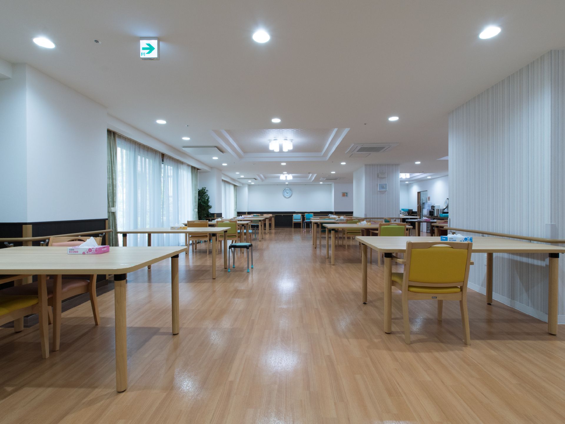 施設利用者が集まる広い部屋である。大き目のテーブルや椅子が数脚、観葉植物などが設置されている。また壁には手すりが取り付けられている。