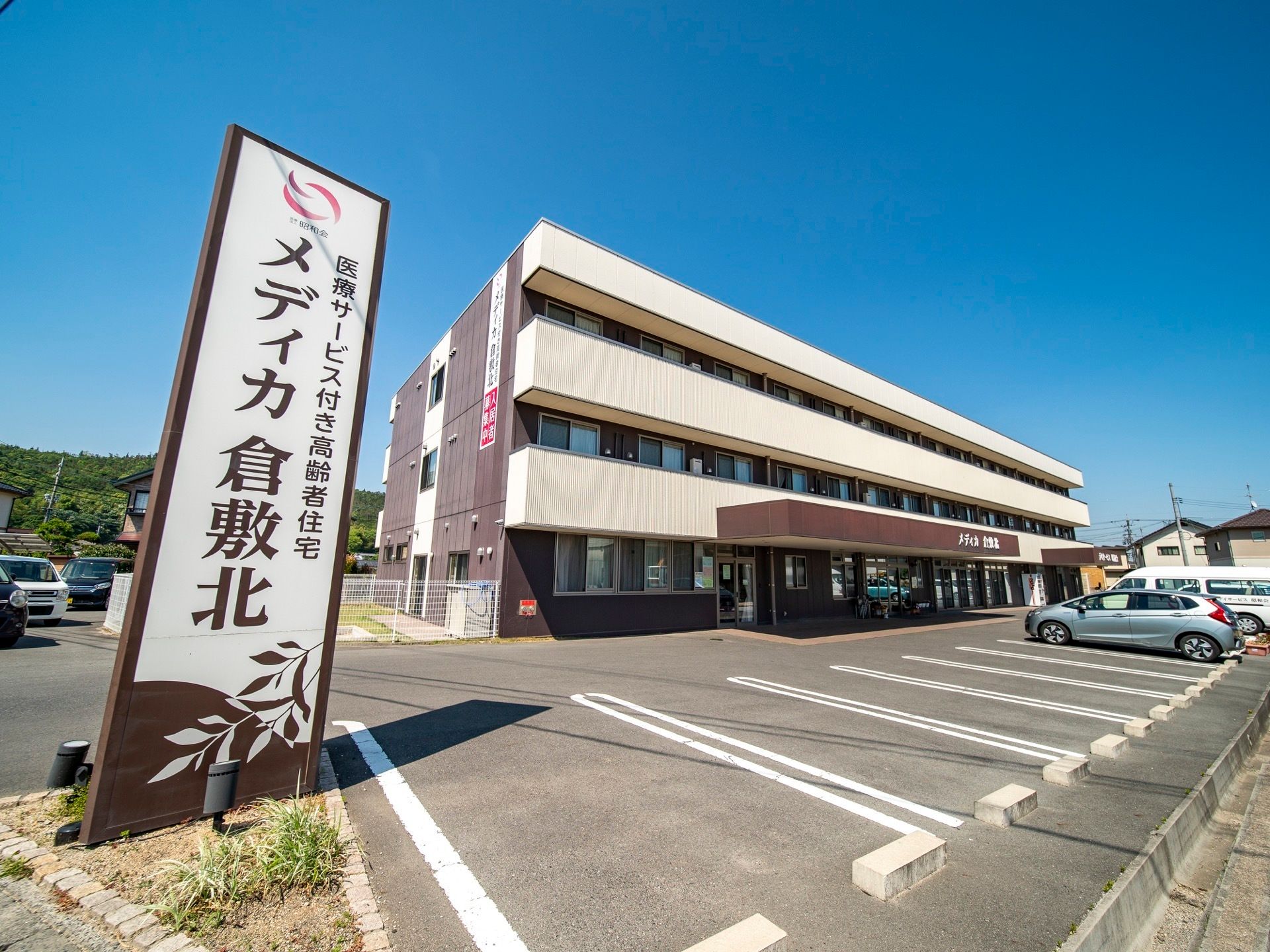 ３階建ての大きな建物の正面が駐車場になっている。地面は舗装され、駐車スペースを示す白線が引かれている。施設名が書かれた看板が立っている。