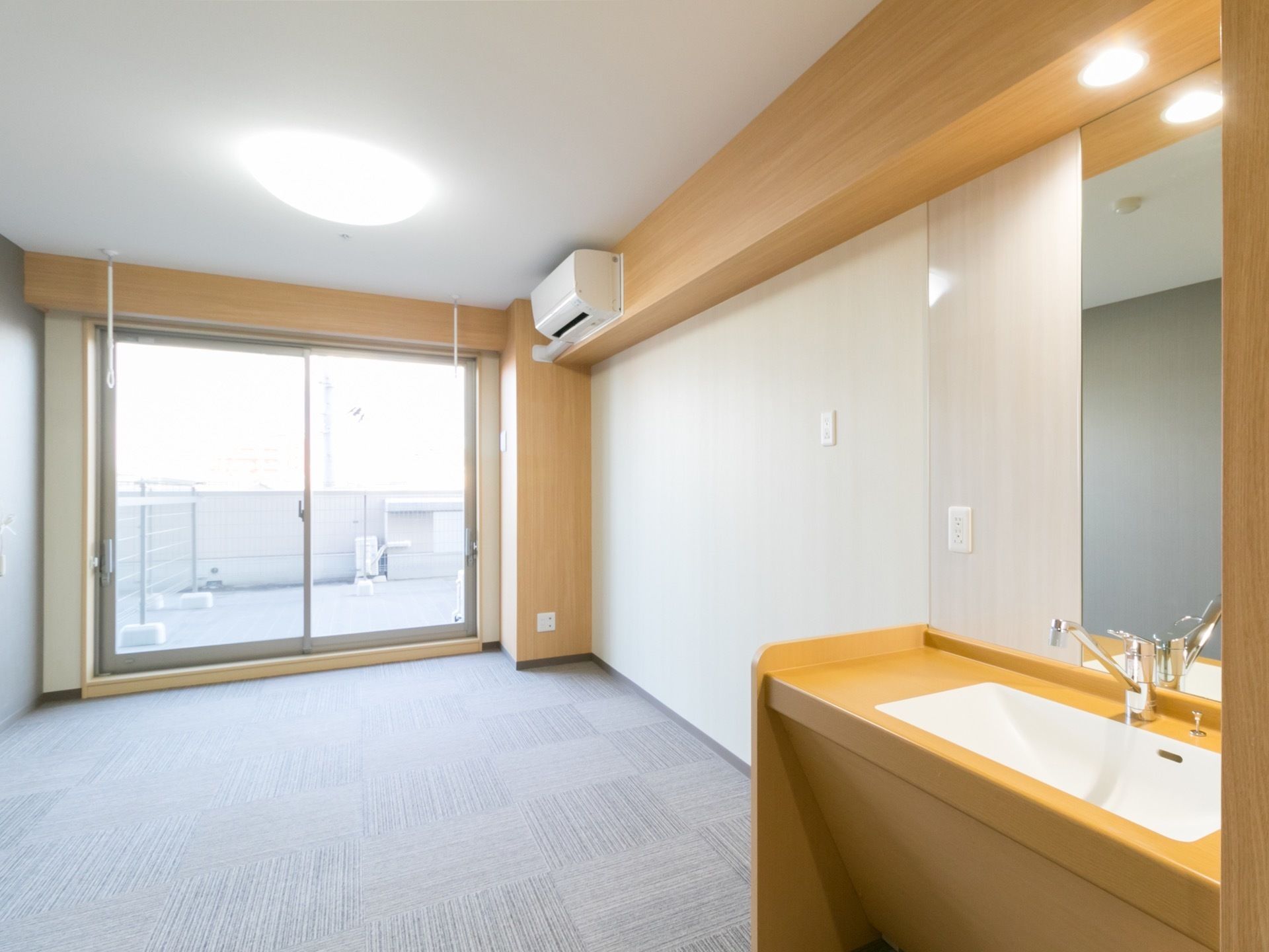 居室には洗面台、エアコンが設置されている。床にはテイルカーペットが全面に敷かれている。広いベランダもある。