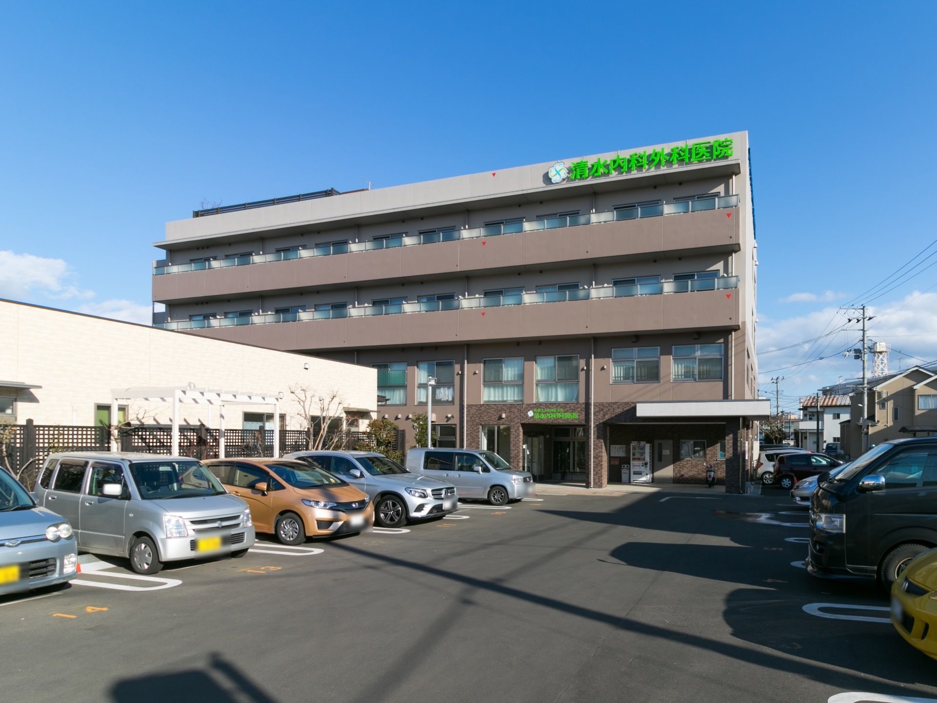施設外観の様子。4階建ての建物の前には駐車場がある。右隣りには民家があり、施設の外壁は、ブラウンを基調とした落ち着きのある色が使われている。