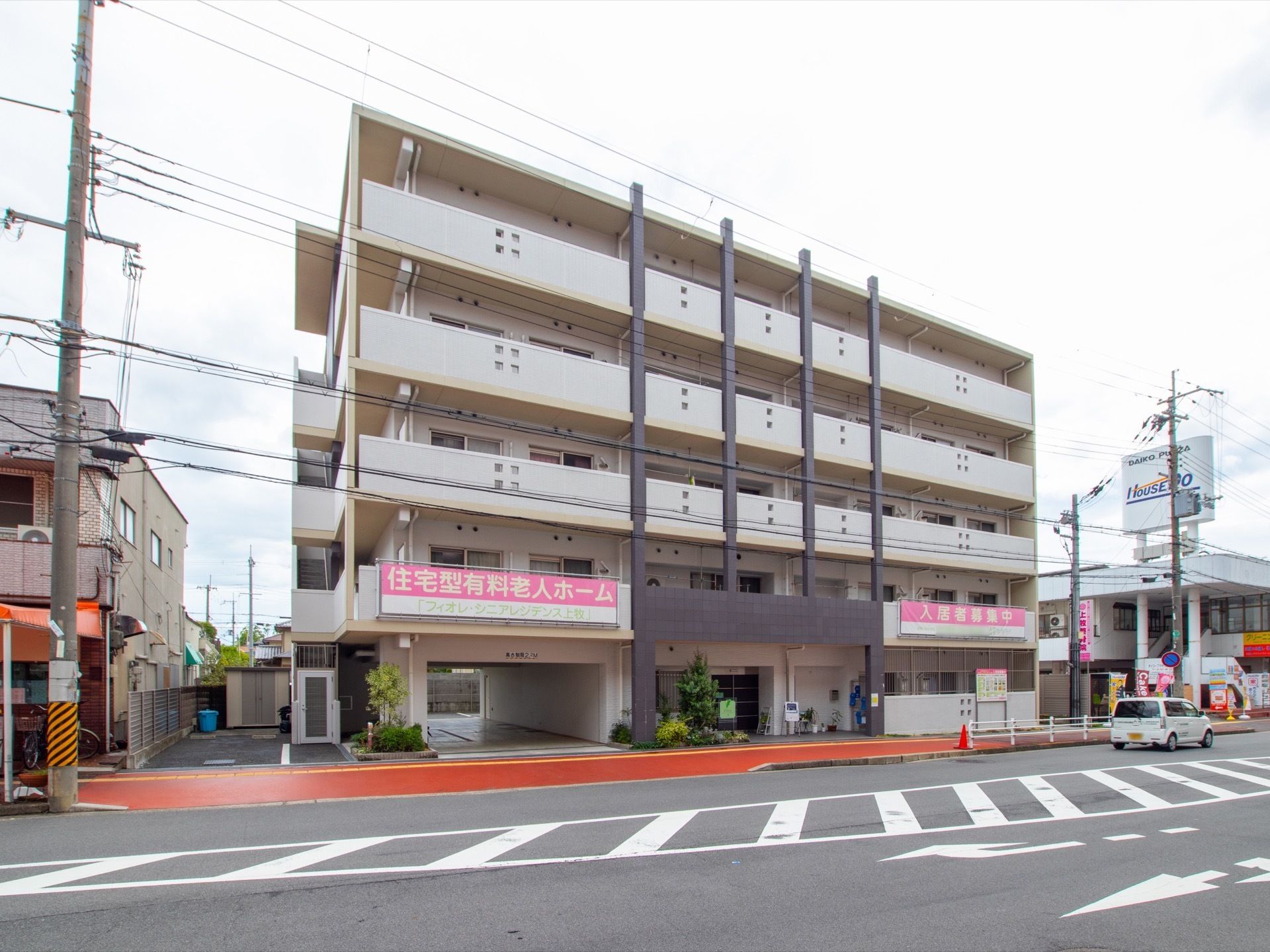 ５階建てのマンションのような白い建物が建っている。施設前の歩道はオレンジ色に塗られ、車道を１台の軽自動車が走っている。