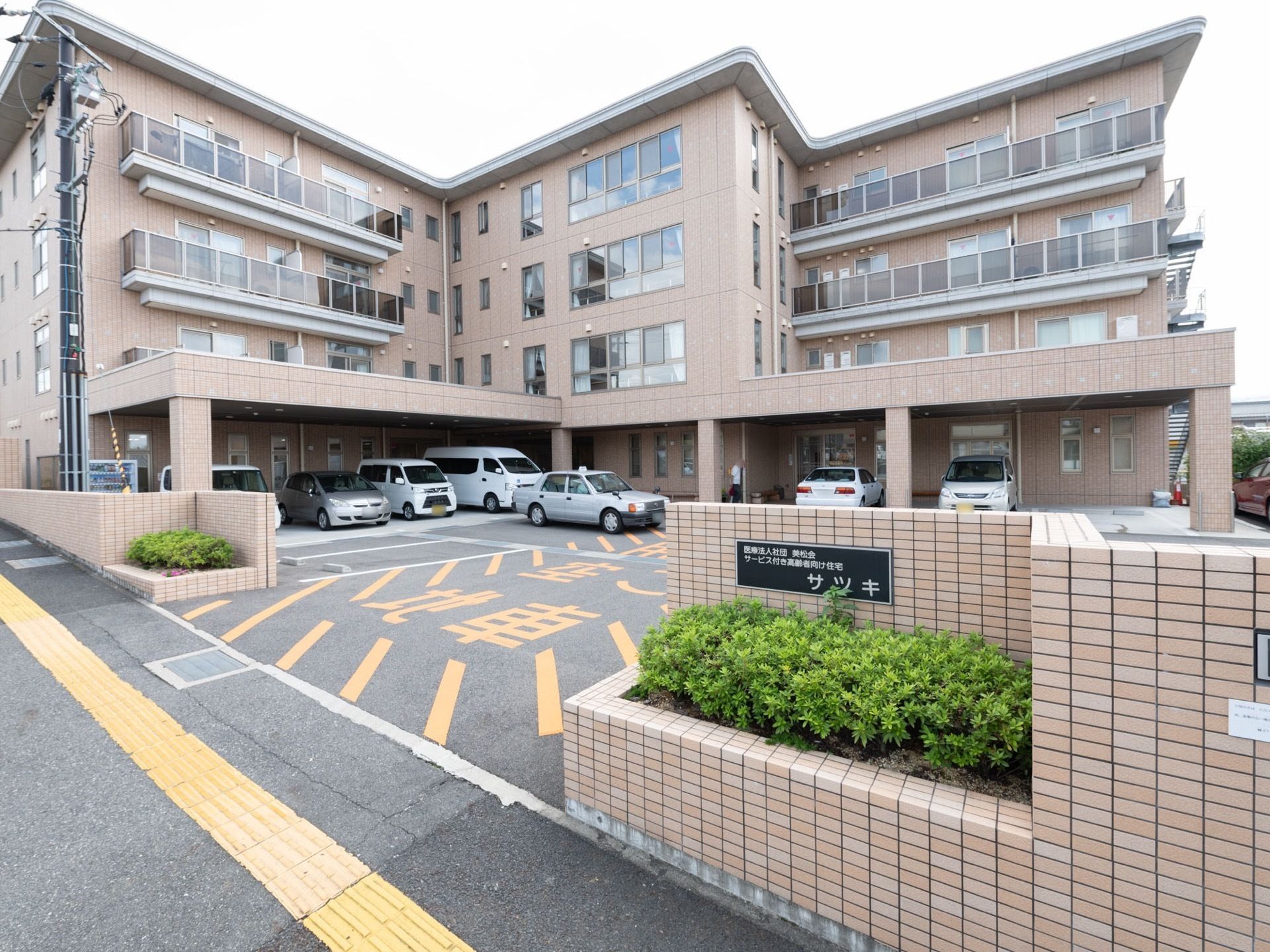 薄い茶色やベージュのタイルが張られた建物が建っている。１階部分には屋根付きの駐車場があり、車が停まっている。