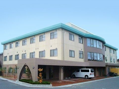 ベージュとブラウンの外壁に緑色の屋根のホームである。建物の前面を覆うように、長い屋根が付いており駐車場の一部も屋根の下にある。