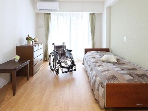 フローリングの床。ベランダに続く大きな窓がついている。ベッド、エアコンが完備されている。車椅子でも広々と利用できる。