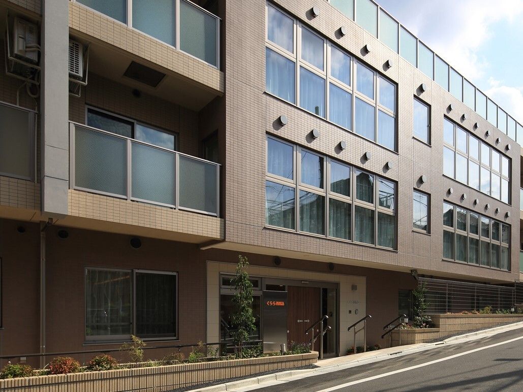 道路に面した４階建ての建物。入口は道路から少し下がっている。階段には手すりがついている。各居室にはベランダがある。