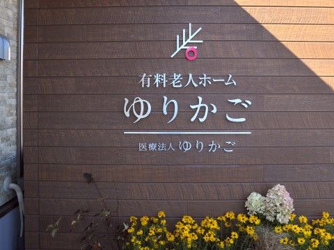 施設ロゴマーク、施設種別、施設名称、医療法人名が、花壇上のこげ茶の壁にシルバー材の文字板で設置されている。