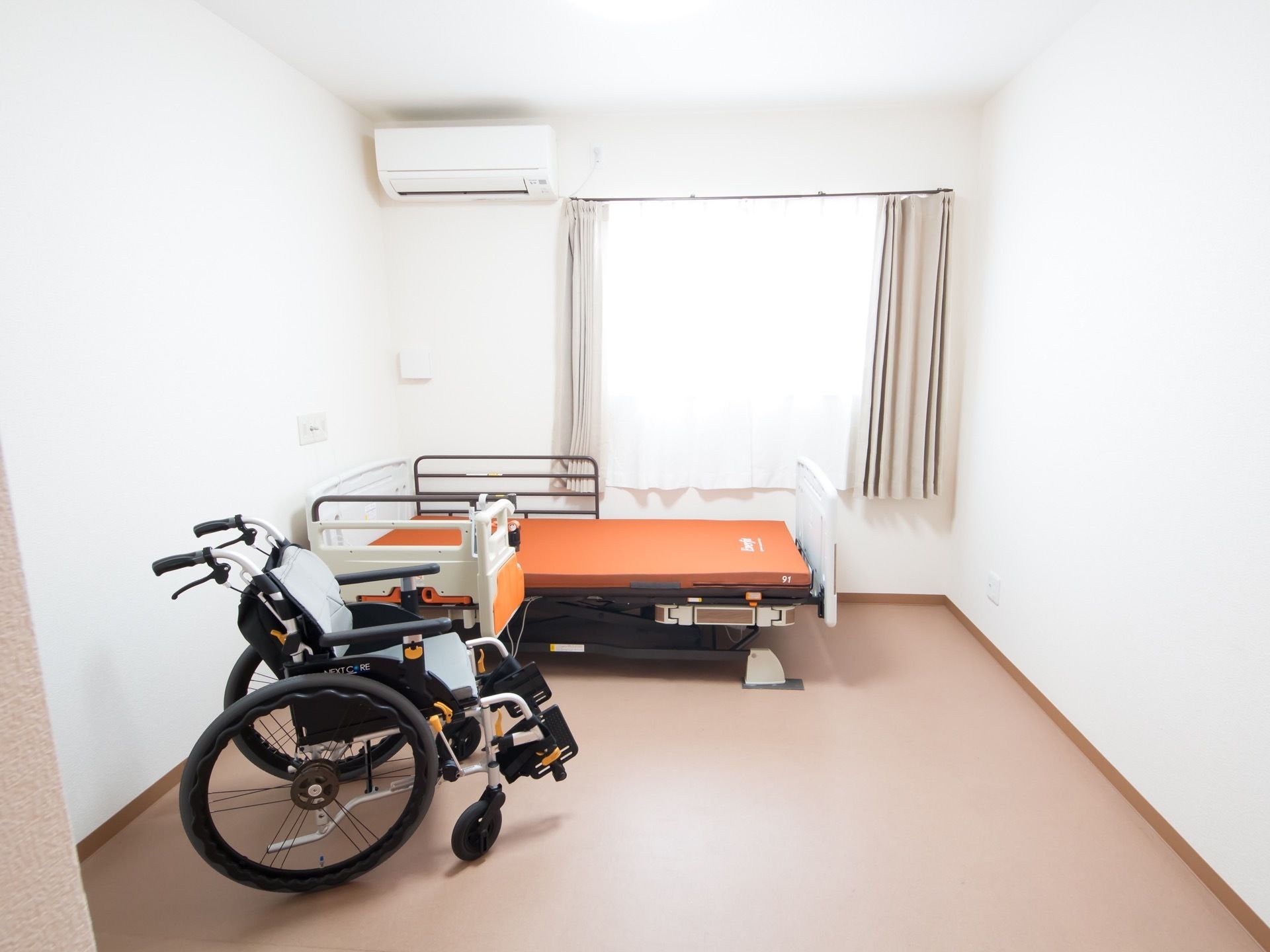 白色の壁のベージュ色の床の部屋で、壁にエアコンが備わっている。室内に介護ベッドと車椅子が置かれている。