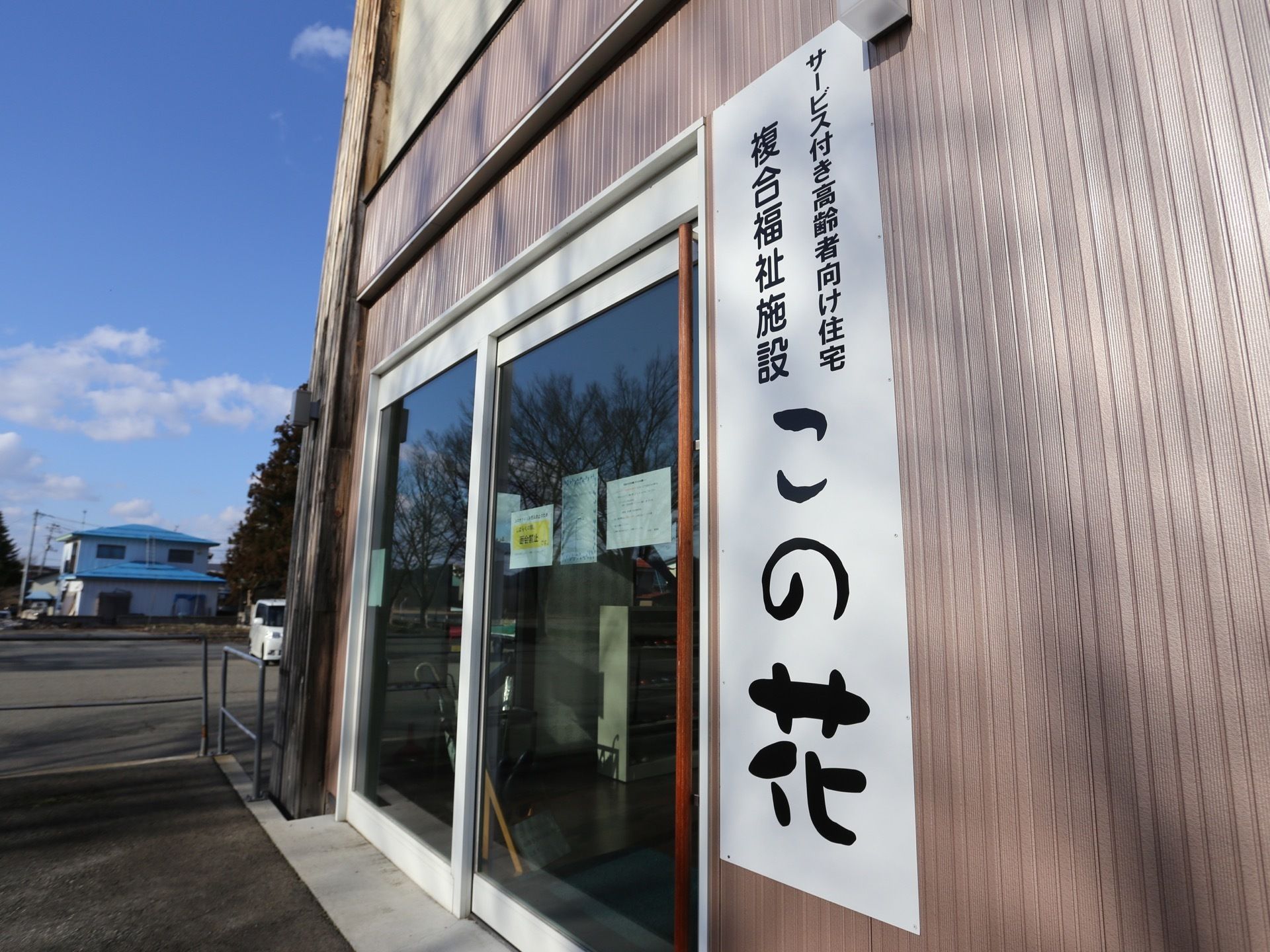 施設入口には、ガラスサッシの扉が設置されている。横の壁面には、施設名称と種別が記載された縦長のプレートが付けられている。