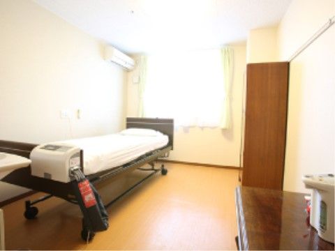キャスター付き介護ベッドがありポットが置かれた棚や収納棚が置かれている。部屋にはエアコンもあり快適に過ごせる。