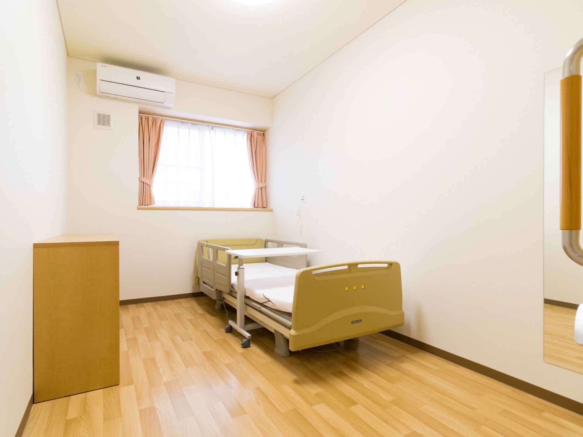 奥に長い部屋である。腰高窓にピンクのカーテンがかかっている。窓の上部にエアコンがある。壁際に手すりのあるベッドがある。