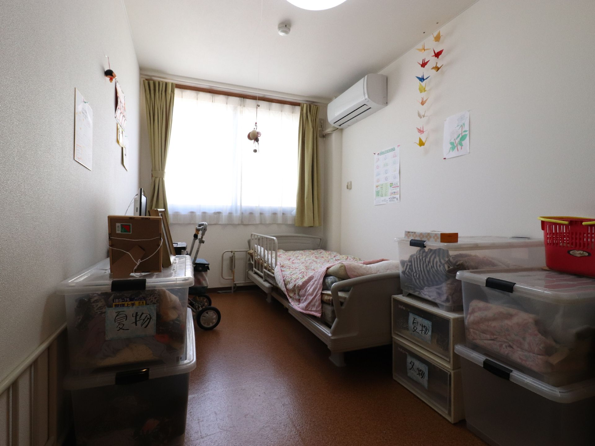 居室には、介護用ベッドやエアコンが設置されており、複数の衣装ケースや歩行車が置かれている。折り鶴が飾られている。