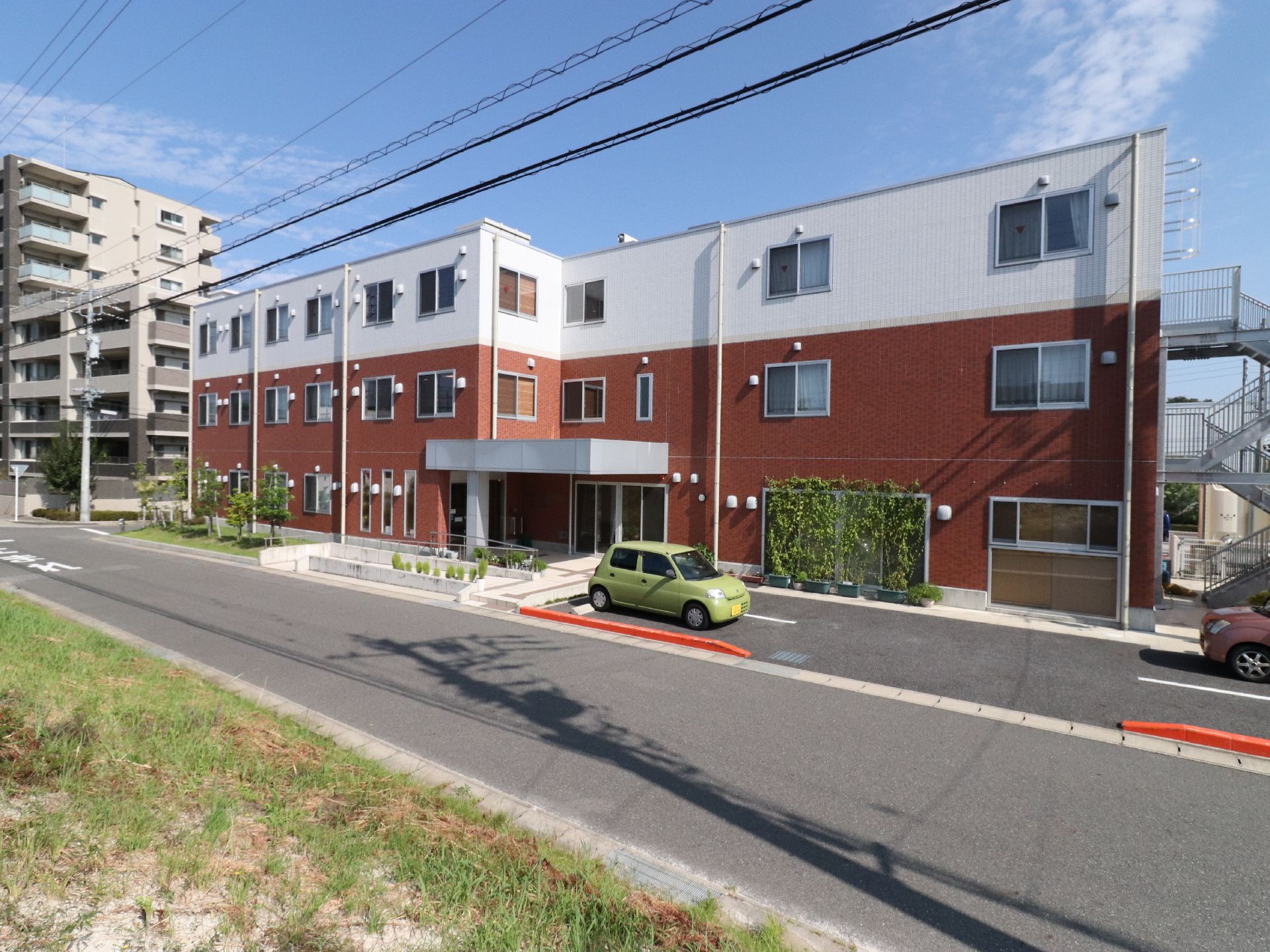 建物が正面に建てられている。そこの左側には木々が植えられている場所がある。左側にはマンションが建っている。