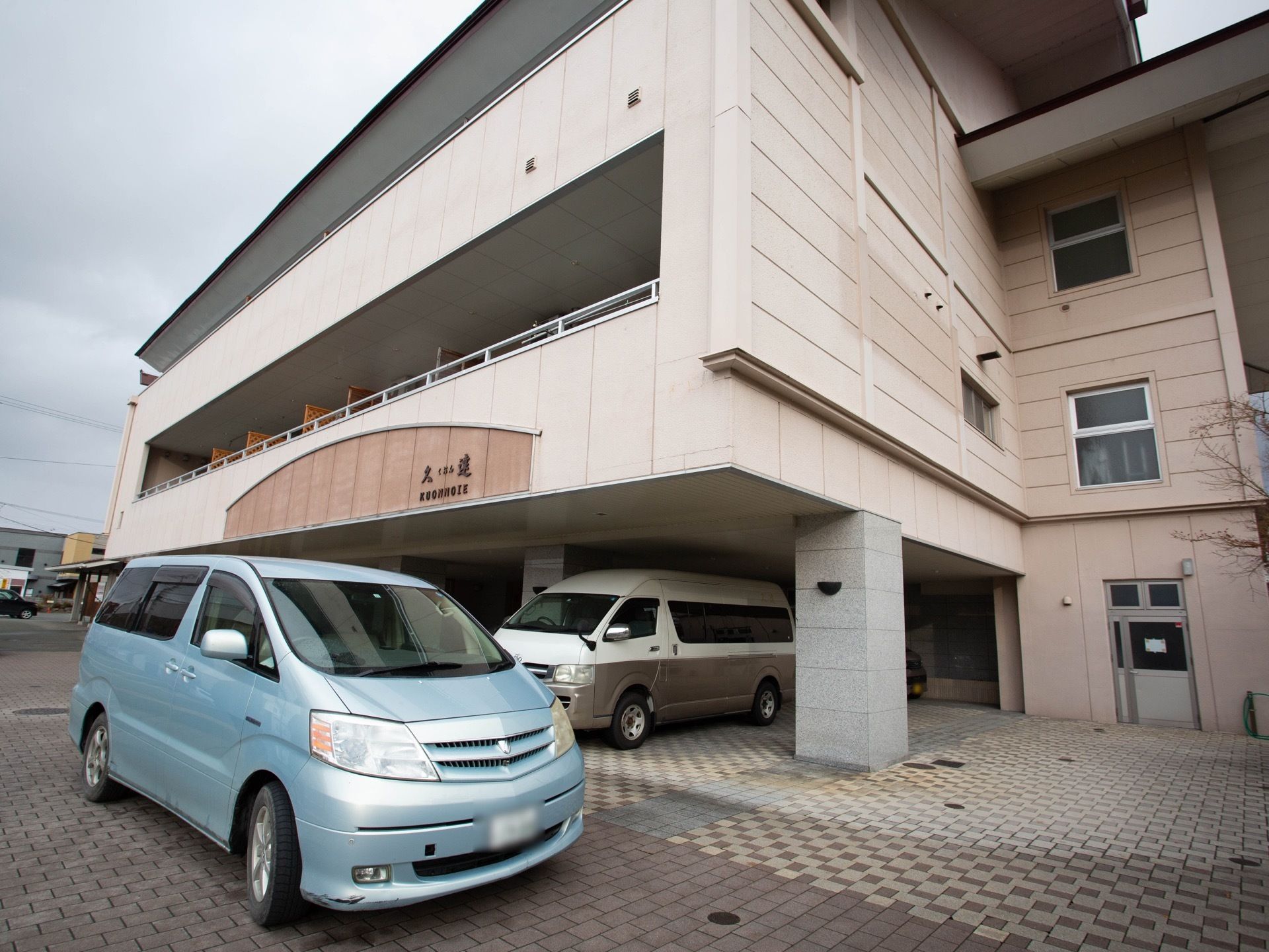 １階部分に駐車スペースがあり、車が停まっている。建物はベージュの外装で、敷地はレンガが敷き詰められている。