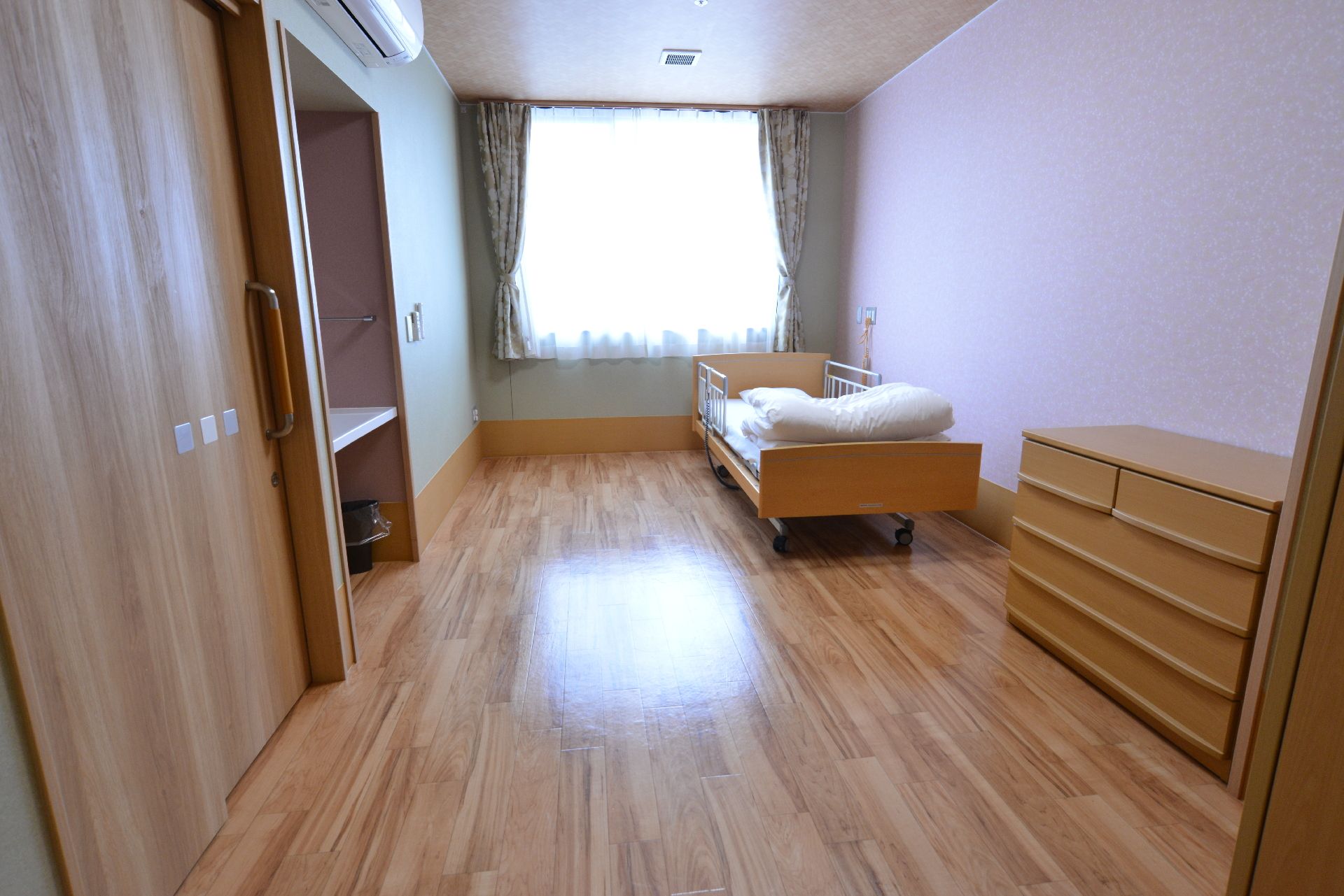 部屋のデザインは全体的にライトブラウンでまとめられている。奥の窓から光が入って明るい。壁にはエアコンも付いている。