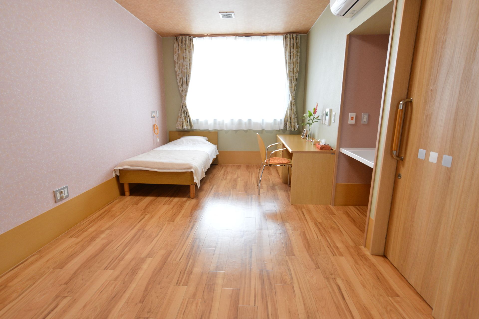 部屋には、ベッドや机とイスが置いてある。家具も床もライトブラウンの色でまとめられている。机の上には花などが飾ってある。