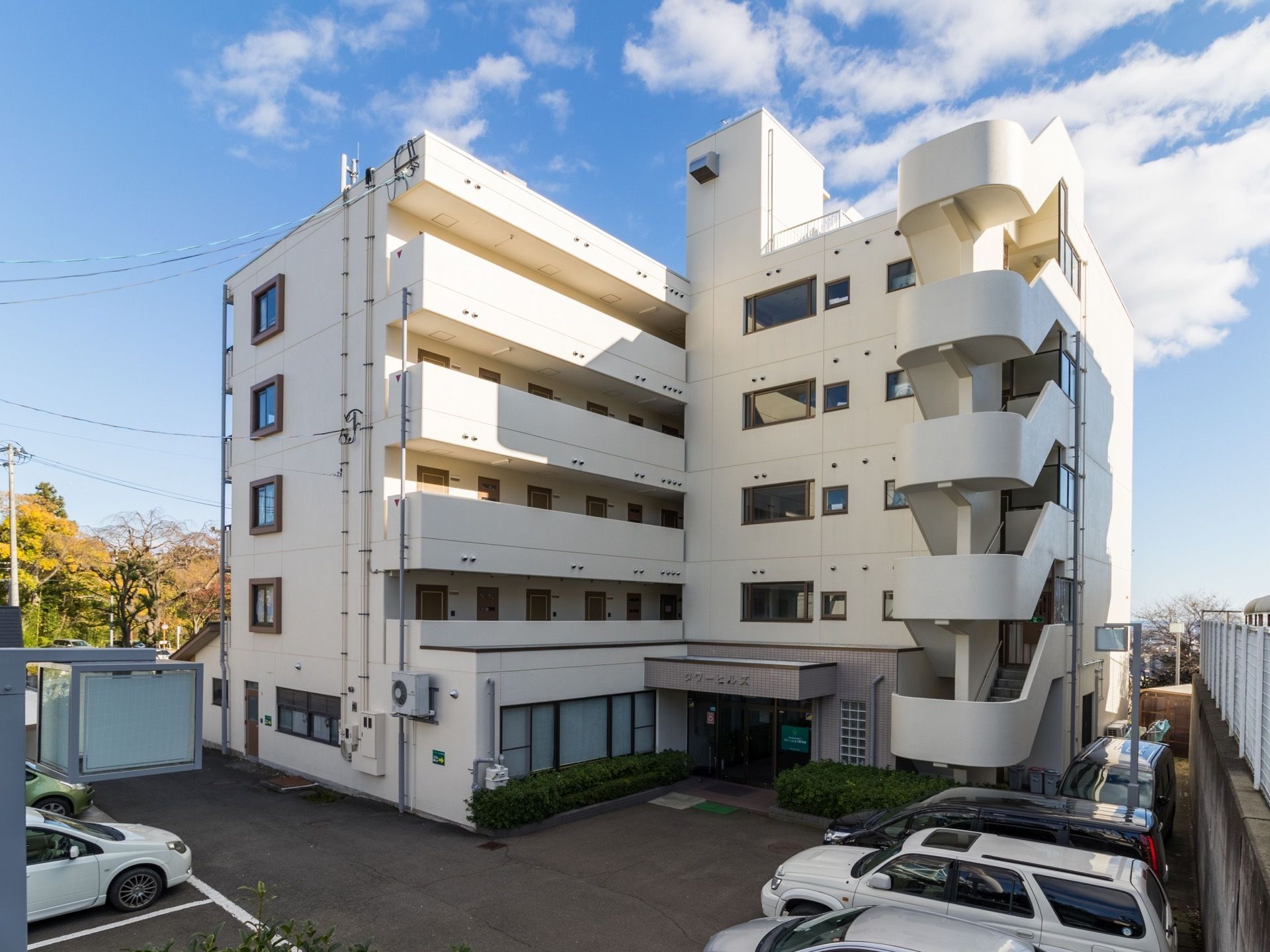 白い大きな建物で、右側には外階段がある。外廊下にはドアがいくつも並んでおり、駐車場には車が数台停まっている。