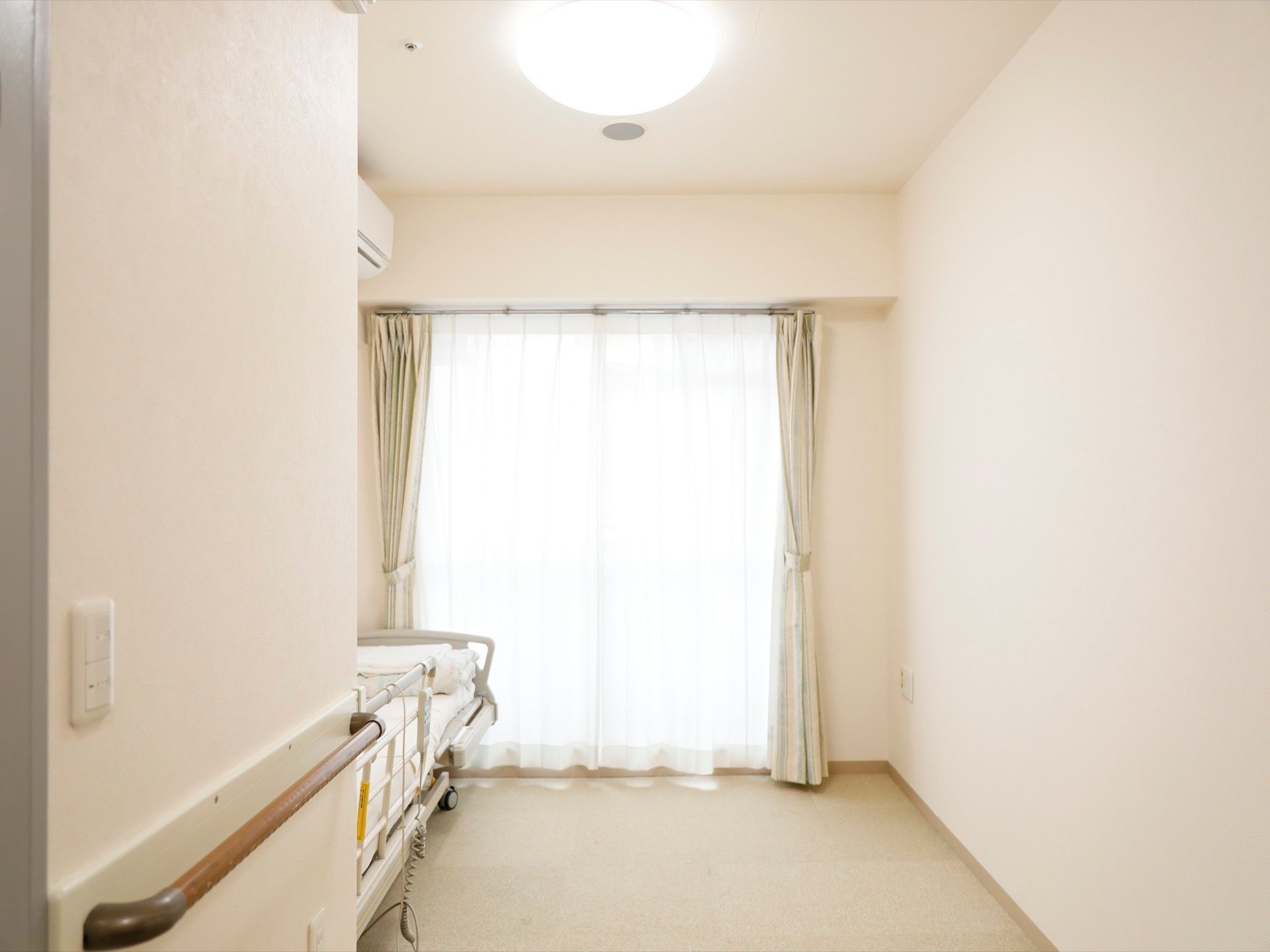 居室は白色で統一されている。介護ベッドやエアコンが設置されている。入口付近には手すりが取りつけられている。