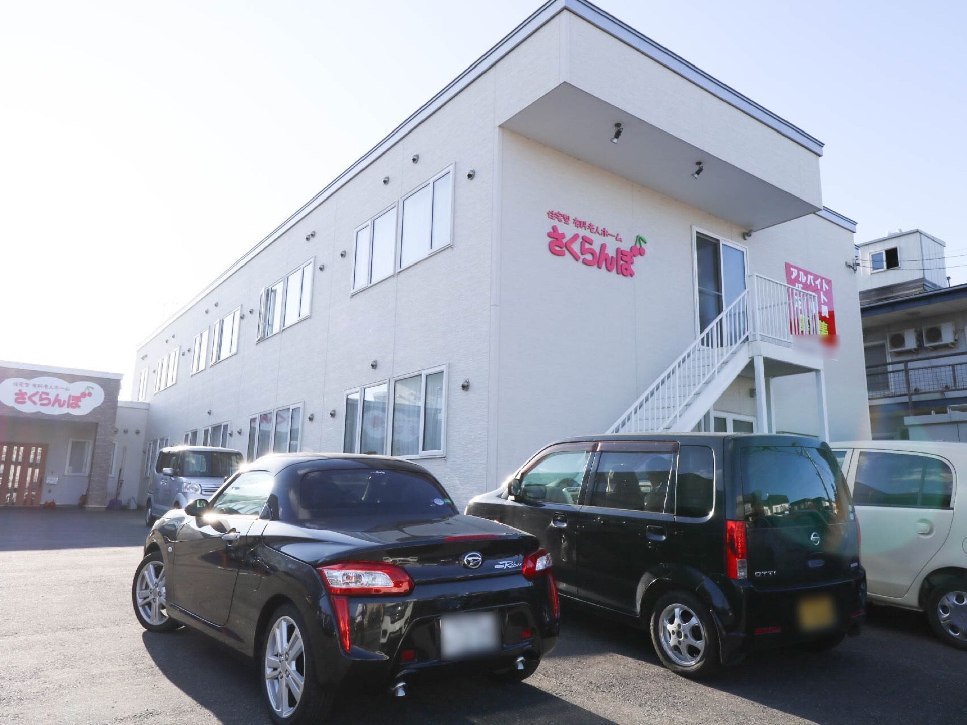 ２階建ての集合住宅が建っている。建物の前はアスファルトで舗装された駐車スペースになっており、車が４台停められている。