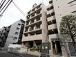 淡いブラウンの外壁の６階建ての建物である。建物の前の植え込みには植物が植えられていて、きれいに手入れされている。