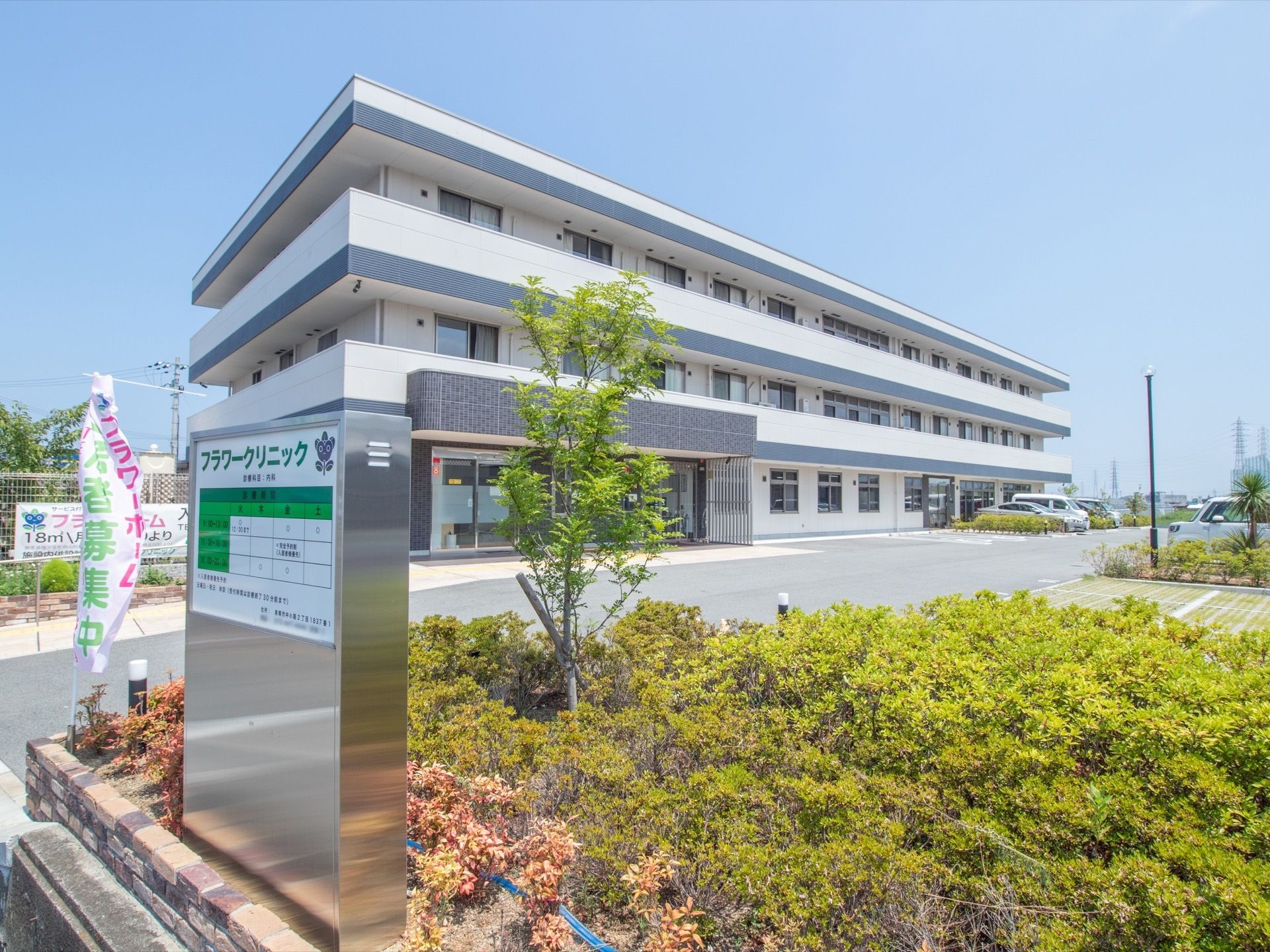 広々とした敷地に、重量鉄骨造の３階建ての施設が建っている。手前の植栽付近に、クリニックの看板が見える。