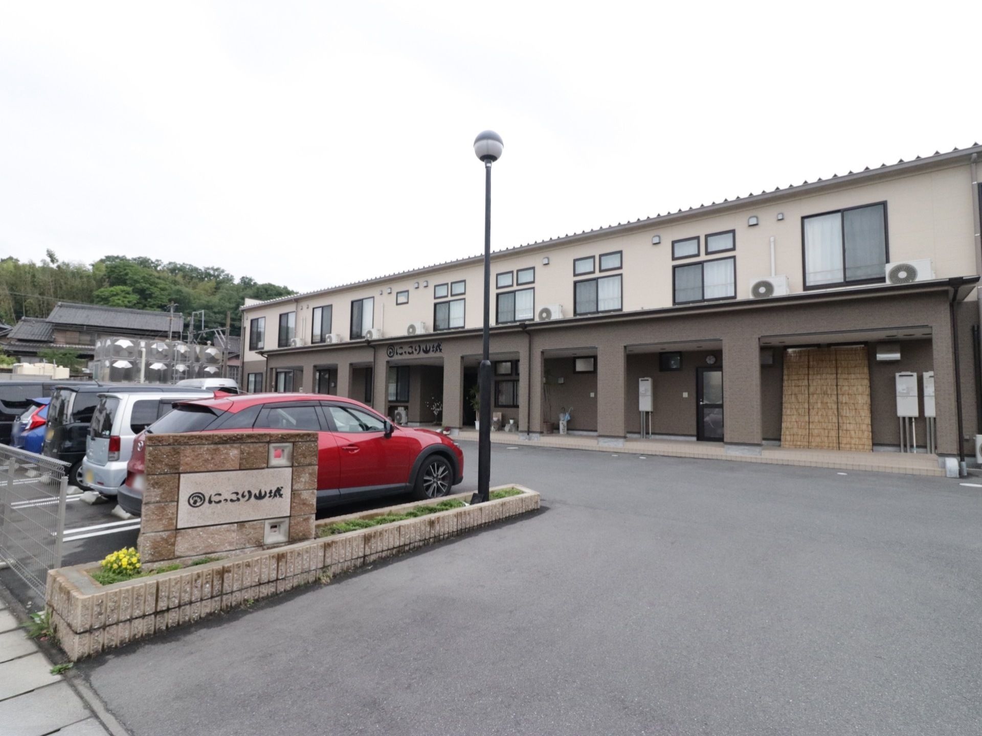 グレーやベージュ色の2階建ての建物である。玄関の前は駐車場になっており、ひさしのすぐそばまで車を寄せることができる。