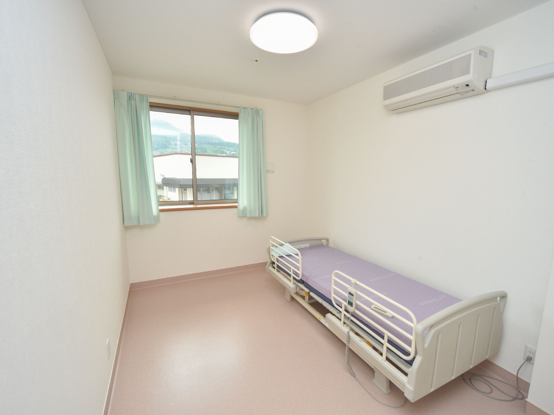 電動の介護ベッドとエアコンが設置されている。窓のすぐ外には向かいの建物があるが、その向こうに山が見える。