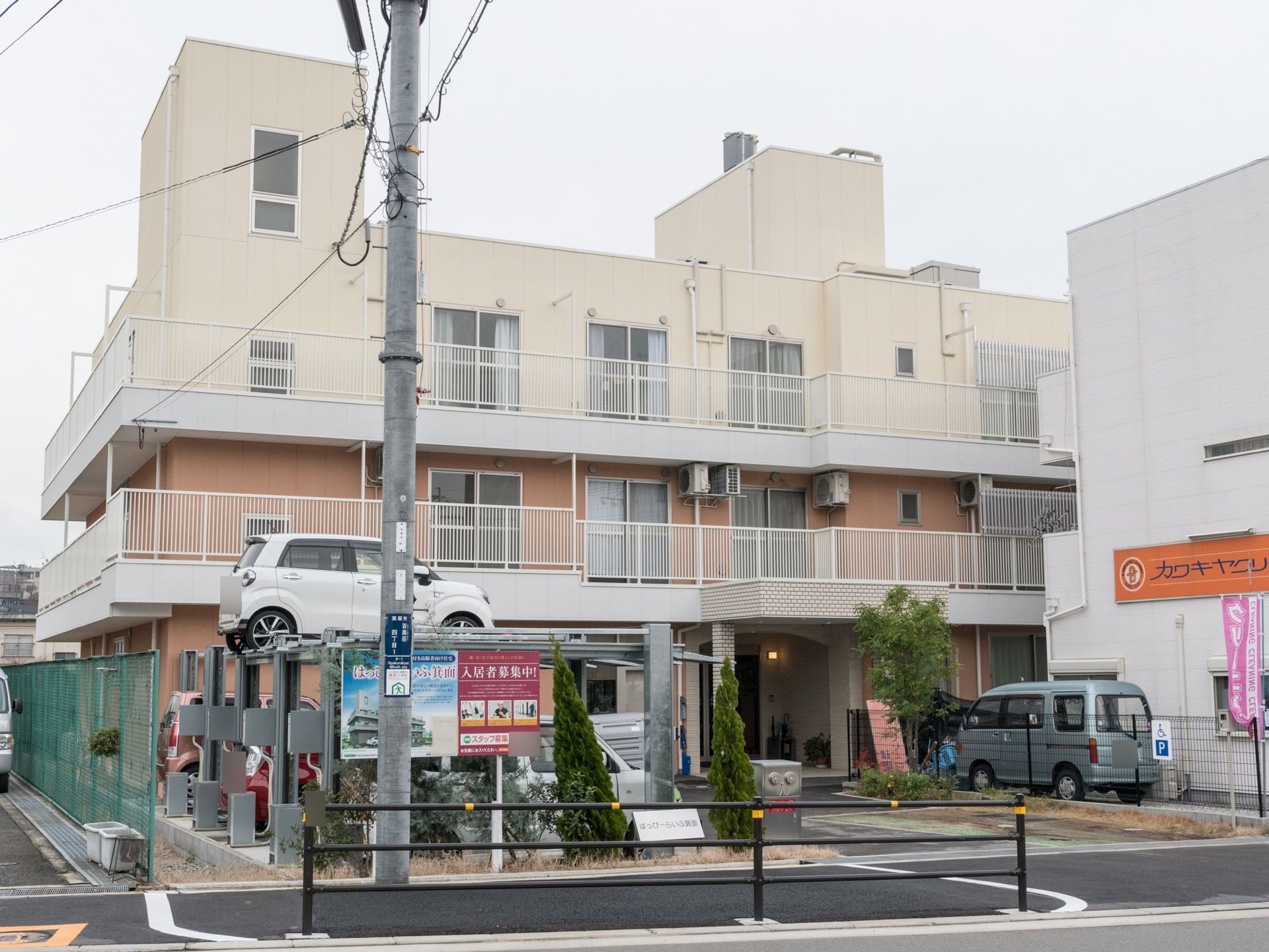 建物には広いベランダが設置されている。施設の前には駐車場がある。入居者募集中の看板が立てられている。