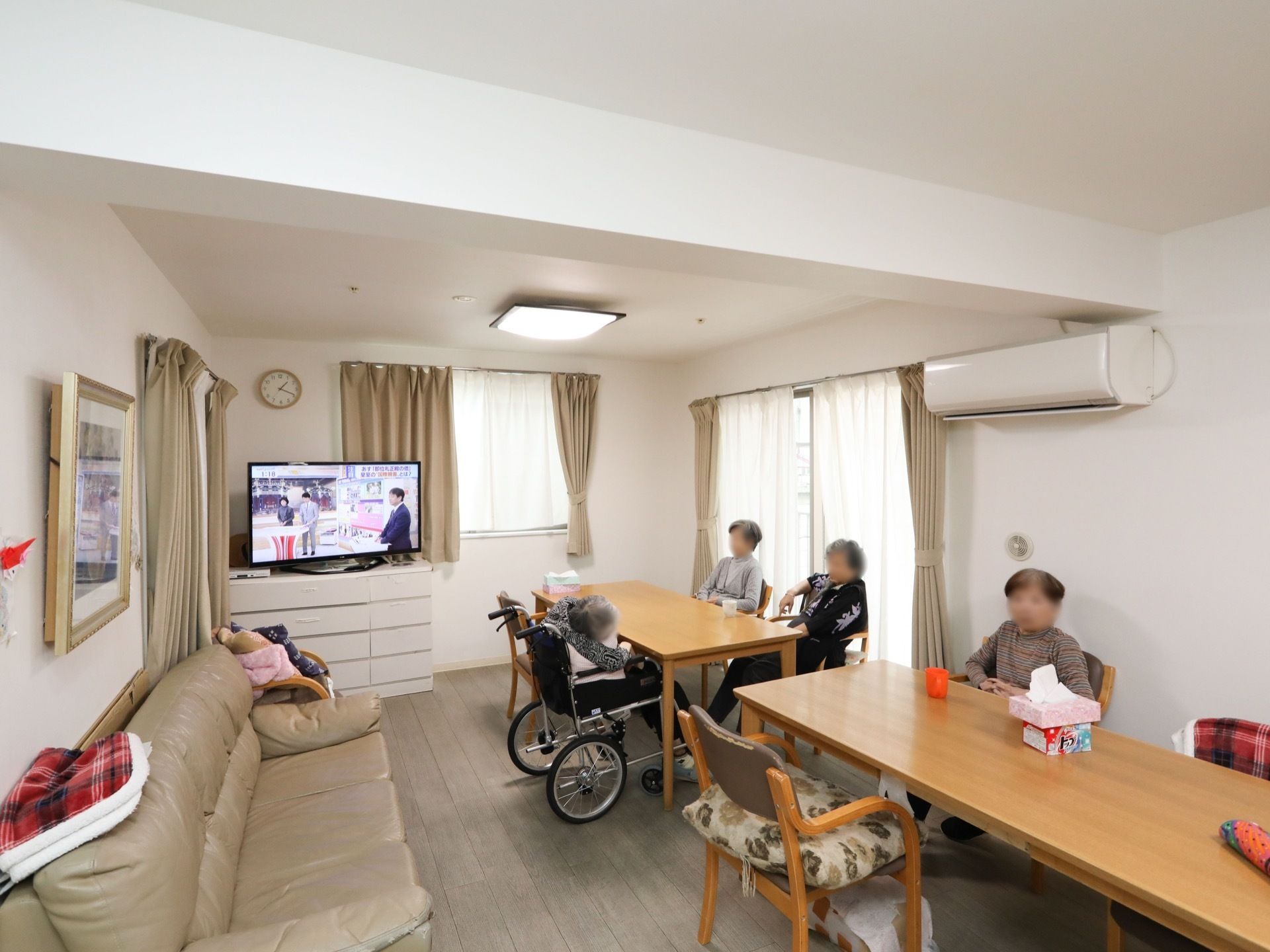 共有スペースにはテーブルが複数あり、入居者様はテレビを観ている。車椅子が通れる間隔に家具が設置されている。