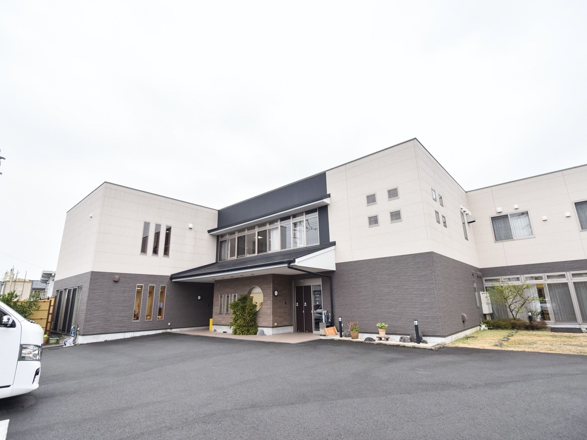 白とグレーのツートンカラーの外壁が印象的な２階建ての建物である。外壁に沿うように樹木やプランターなどが置かれている。