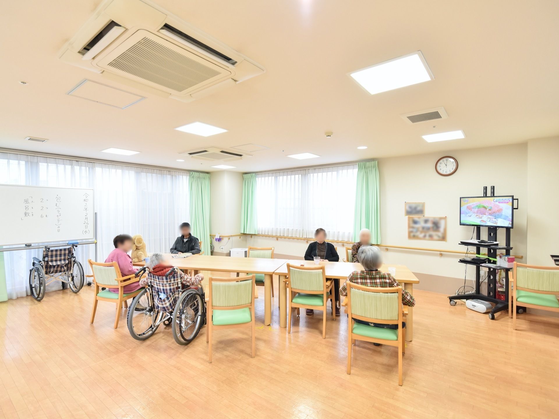 大きな窓が設置された明るい共有スペースである。窓の前にホワイトボードが置かれている。その下には車椅子がある。