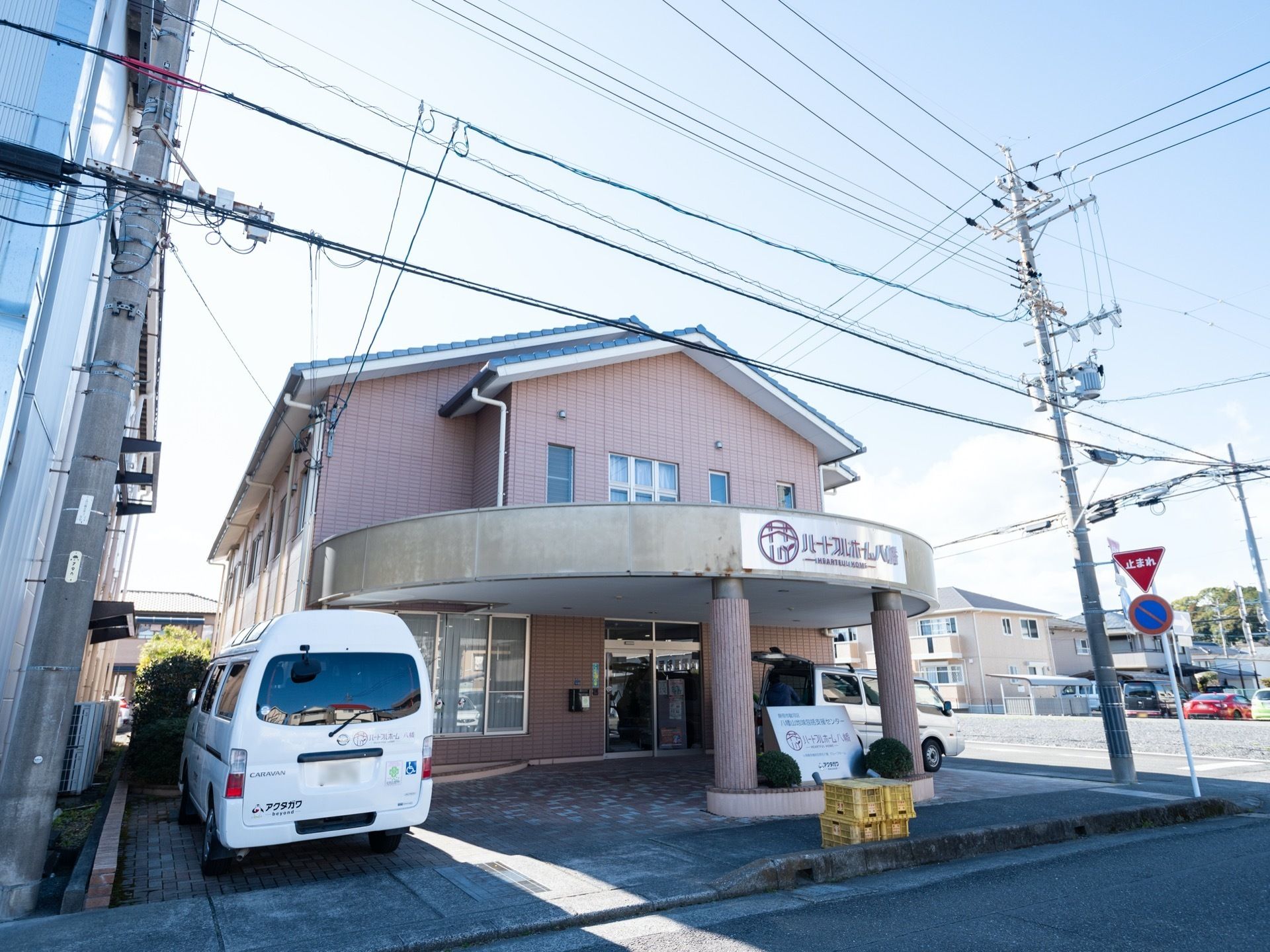 落ち着いた色合いに施された2階建ての建物である。外壁には施設名が記された看板が設置されている。福祉車両が停まっている。