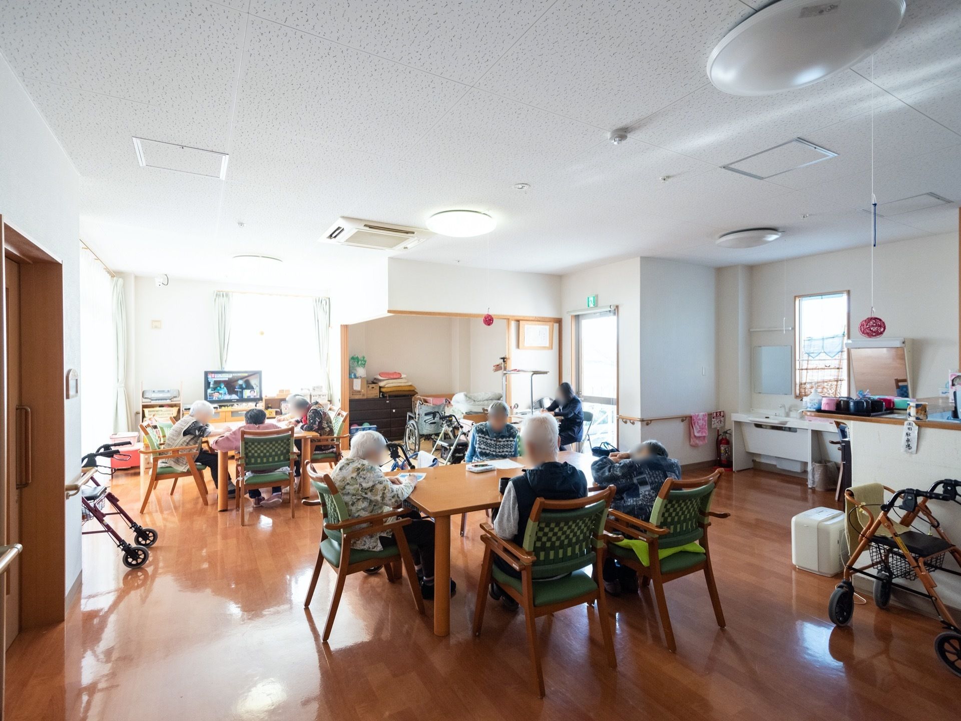 開放的な共有スペースでは入居者様がそれぞれ過ごしており、歩行補助車が用意されている。奥にはキッチンが併設されている。