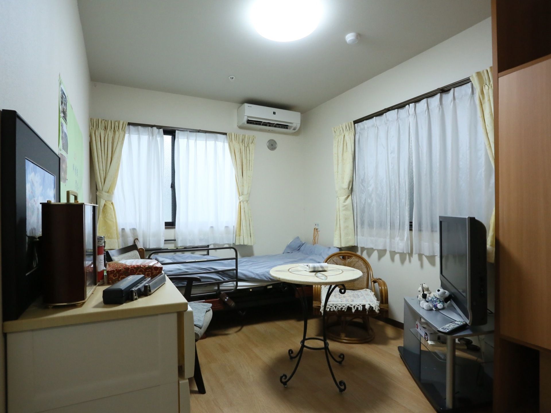 窓が２か所ある。テレビボードの上に液晶テレビが置かれている。タンスの上には小物や写真が飾られている。