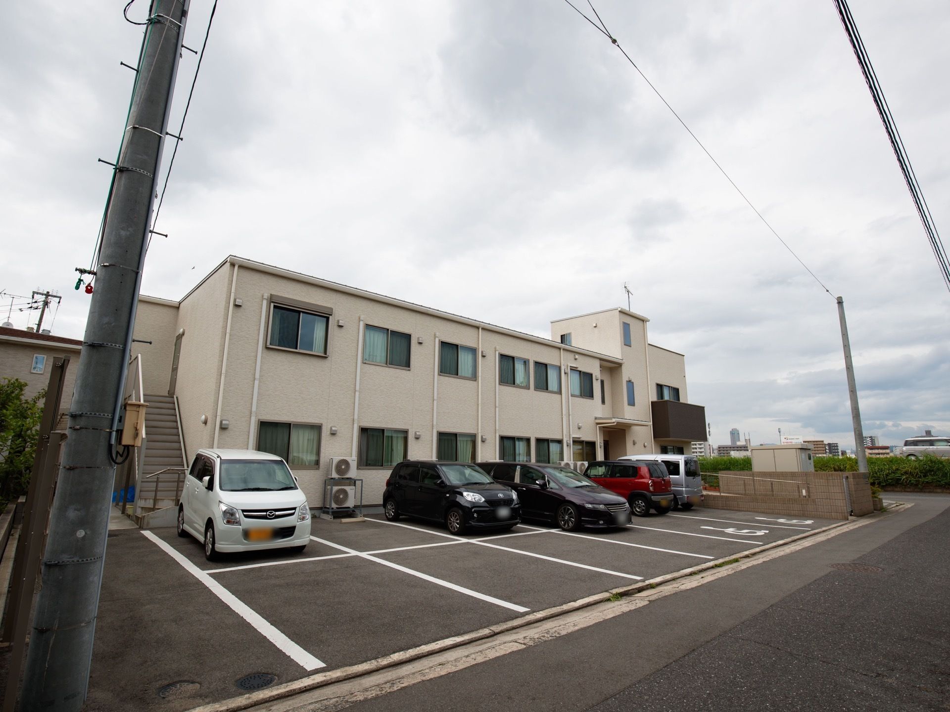 ２階建ての白い建物の裏側に、道路に面した駐車場がある。車が数台停まっている。建物の側面には外付けの非常階段がある。