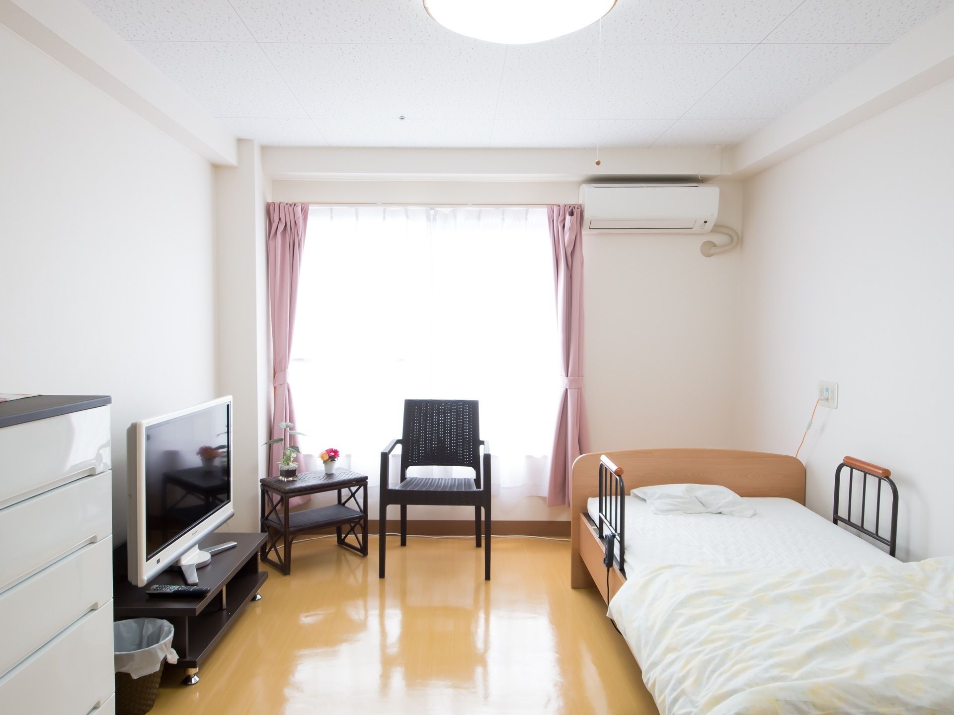 大きな窓のある明るい居室。白い壁と光沢のあるベージュの床があり、明るい雰囲気がある。介護用ベッドとテレビ、椅子、棚が置かれている。