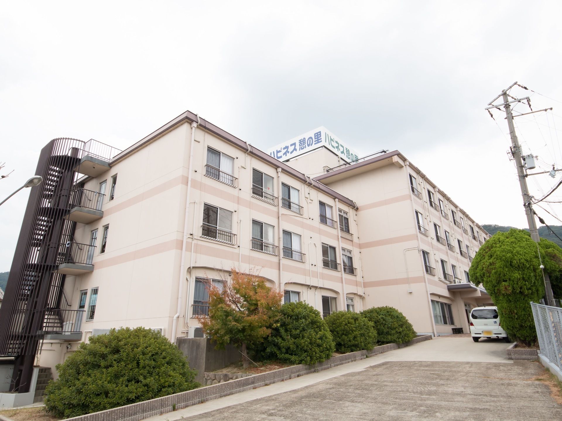 四階建ての二棟続いている大きな建物がある。道の脇には樹木が植えられている。屋上には大きな施設名が書かれた看板が設置されている。