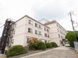 四階建ての二棟続いている大きな建物がある。道の脇には樹木が植えられている。屋上には大きな施設名が書かれた看板が設置されている。
