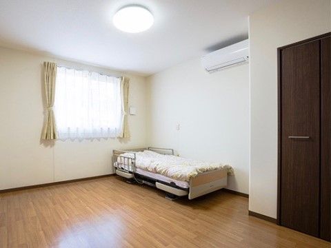 フローリングの広々とした洋室である。手前には収納があり、ベッドのある壁側にはエアコンが設置されている。