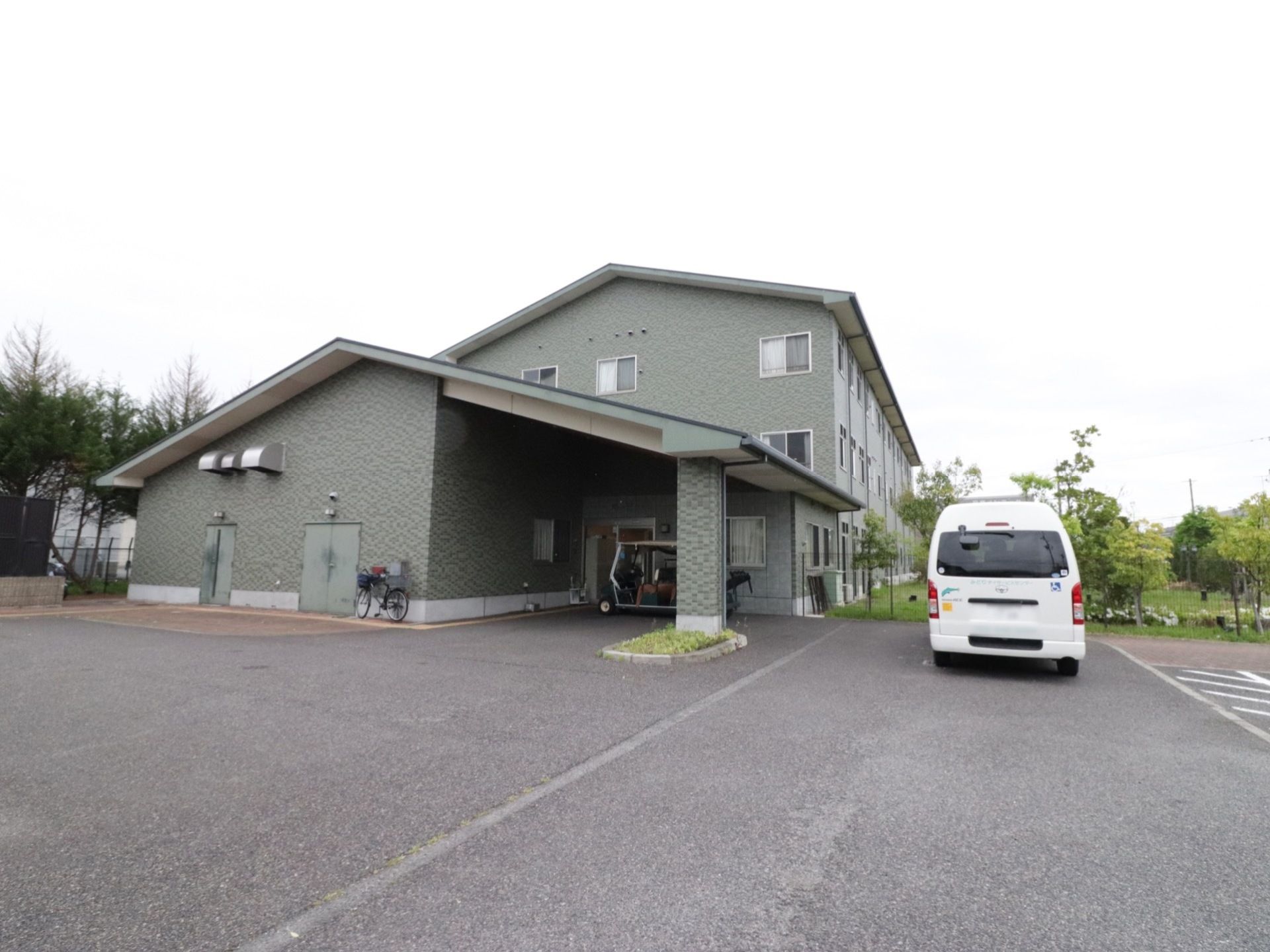 住宅型老人ホームは3階建てになっている。外壁は石造りのように見える。正面玄関の近くに白い車が停められている。車の奥には庭が続いている。