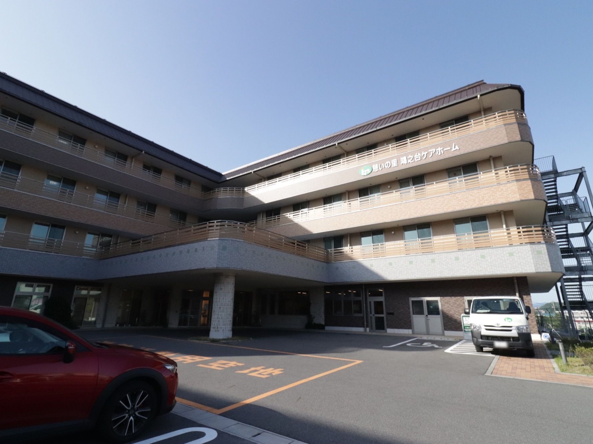 ブラウンを基調とした4階建ての施設の外観。敷地内に駐車場が完備されている。入り口の前には屋根が設置されている。