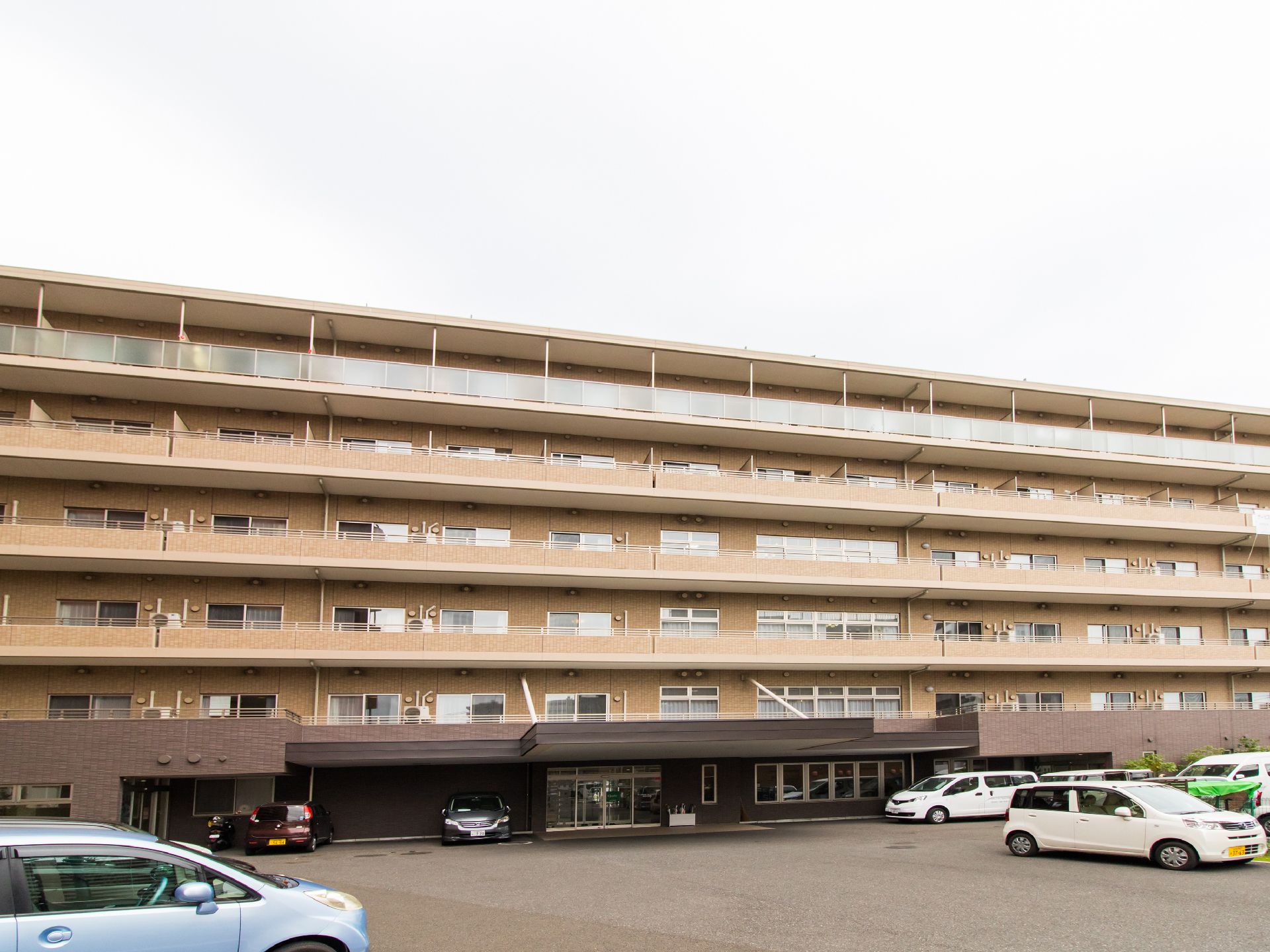 ベージュとブラウンの2色の外壁になっており、建物は6階建てになっている。手前は駐車場になっており、たくさんの車が停まっている。