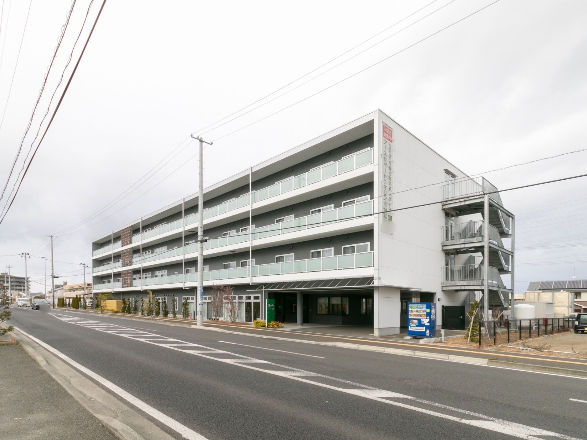 ４階建てのマンションタイプの建物である。向かって右側に、玄関や駐車場入り口がある。角に自動販売機が置かれている。