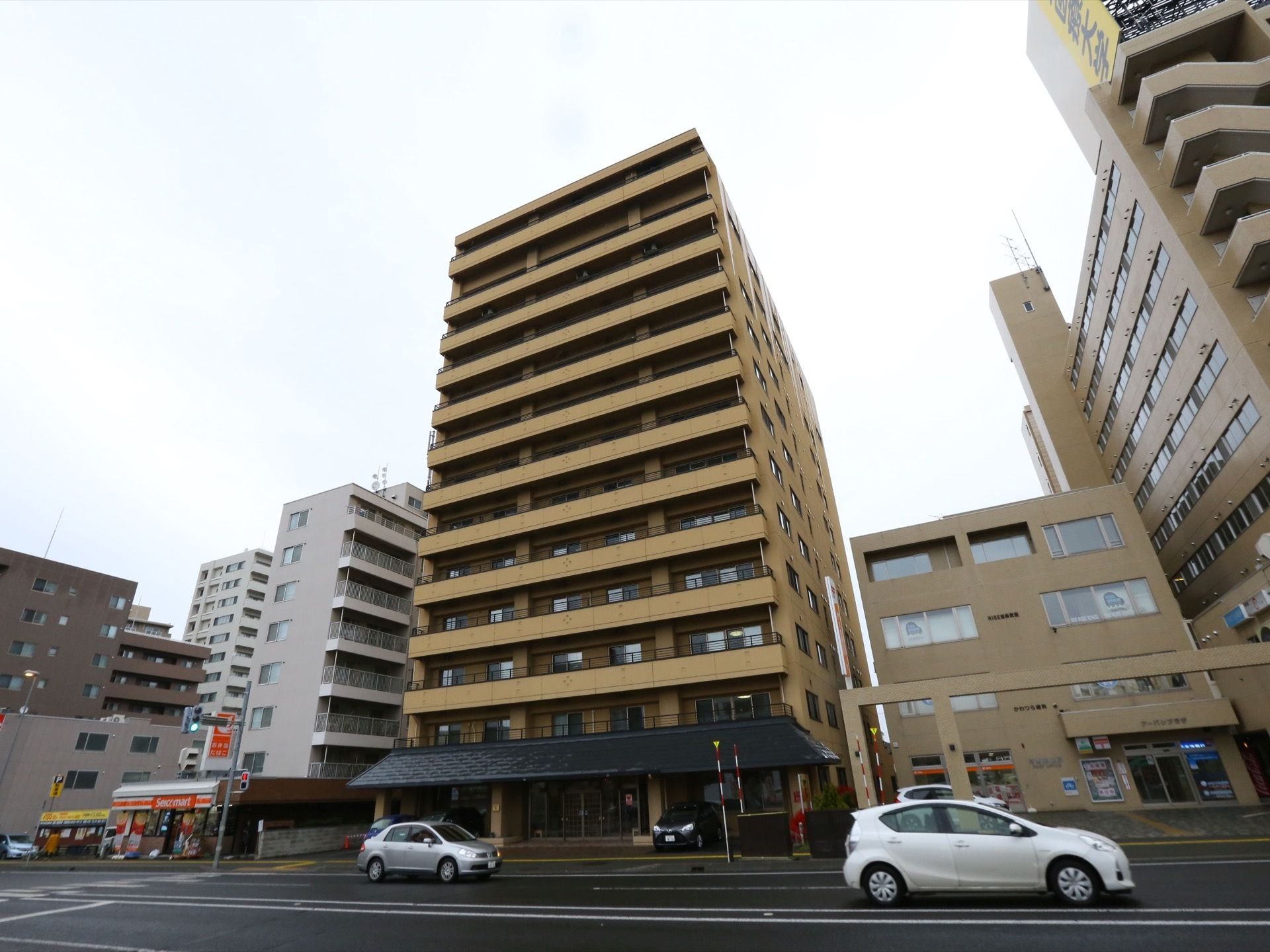 マンションのような外観の大きなグループホーム。広い道路に面しており、隣にはコンビニエンスストアがある。