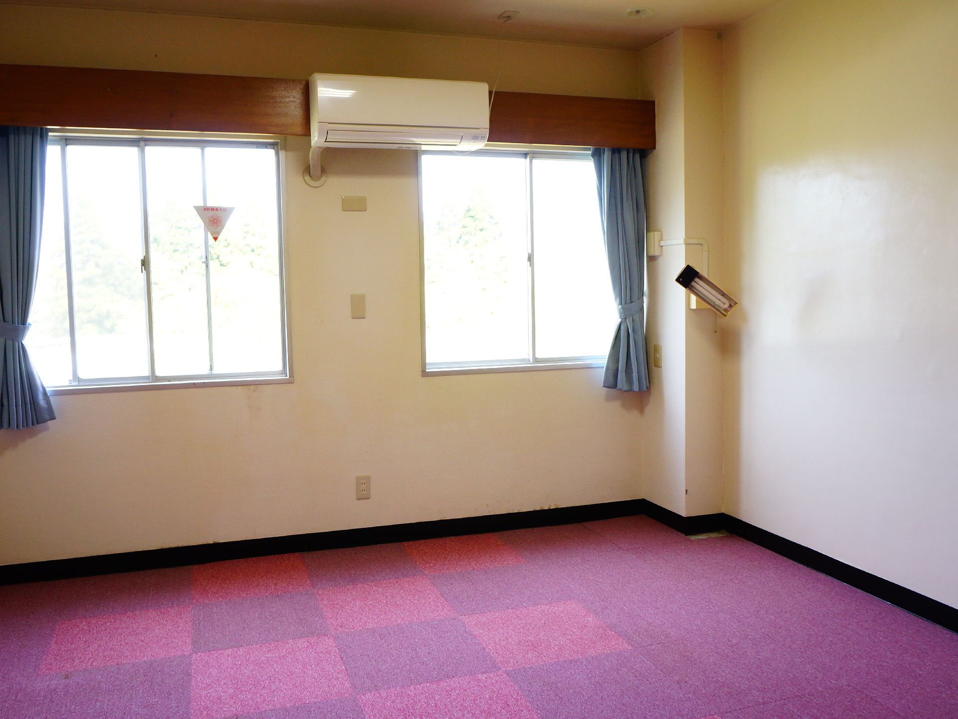 入居者用の居室には淡いブルーのカーテンがかけられており、2つ並んだ窓の中央上部にエアコンが設置されている。