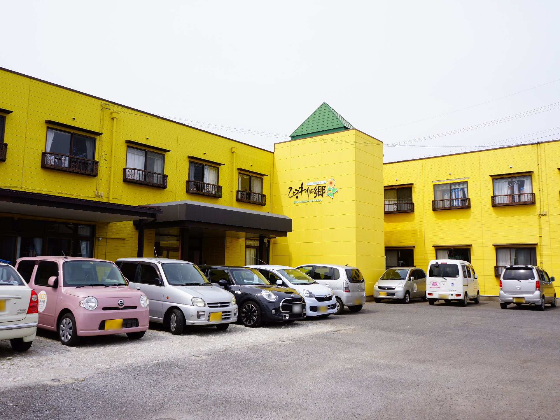 正面に2階建ての黄色い外壁の建物がある。そこの手前には複数の車が横並びに止まっている。正面奥の外壁には施設名が記載されている。